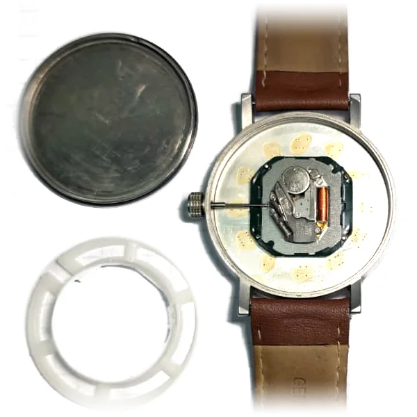 Geöffnete Armbanduhr mit Uhrwerk und Knopfzelle, daneben Gehäuseboden und Distanzring